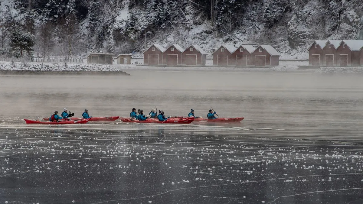 Kayakers paddling across a misty, half-frozen fjord past red boathouses