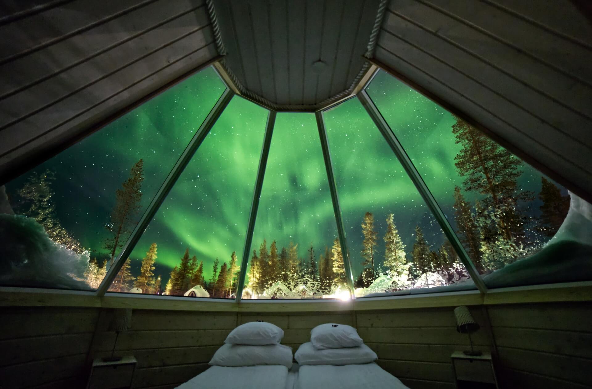 The northern lights seen from the bed beneath a glass-roofed cabin