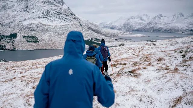 Hikers in blue jackets walking across a snowy hillside above a fjord