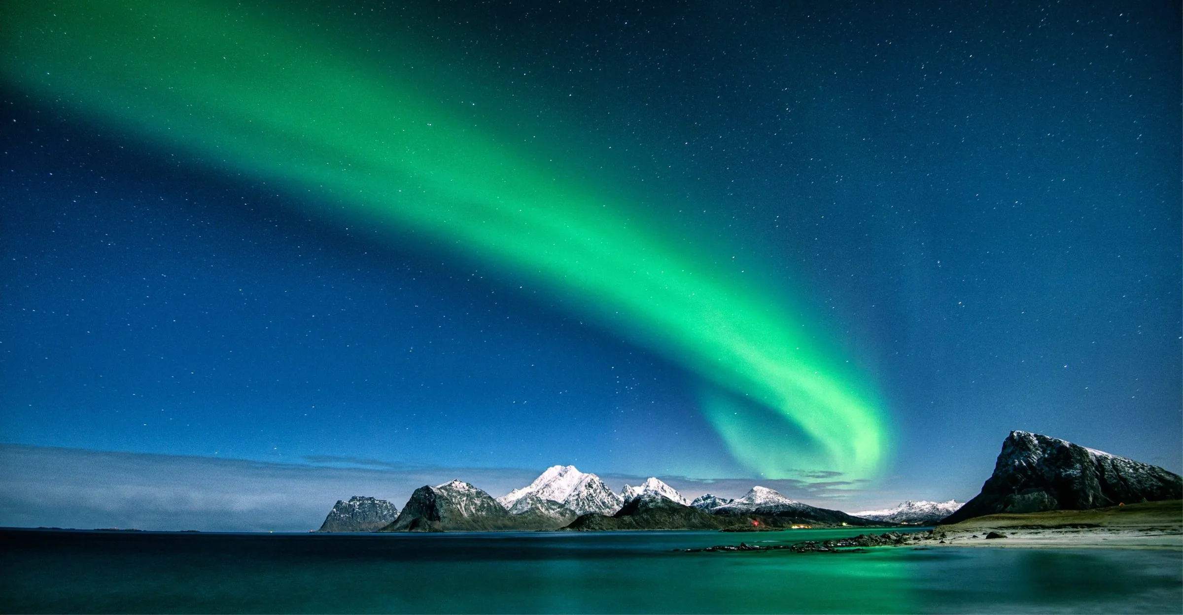 The Northern Lights swirling over snow-capped peaks and a still bay in Lofoten, Norway