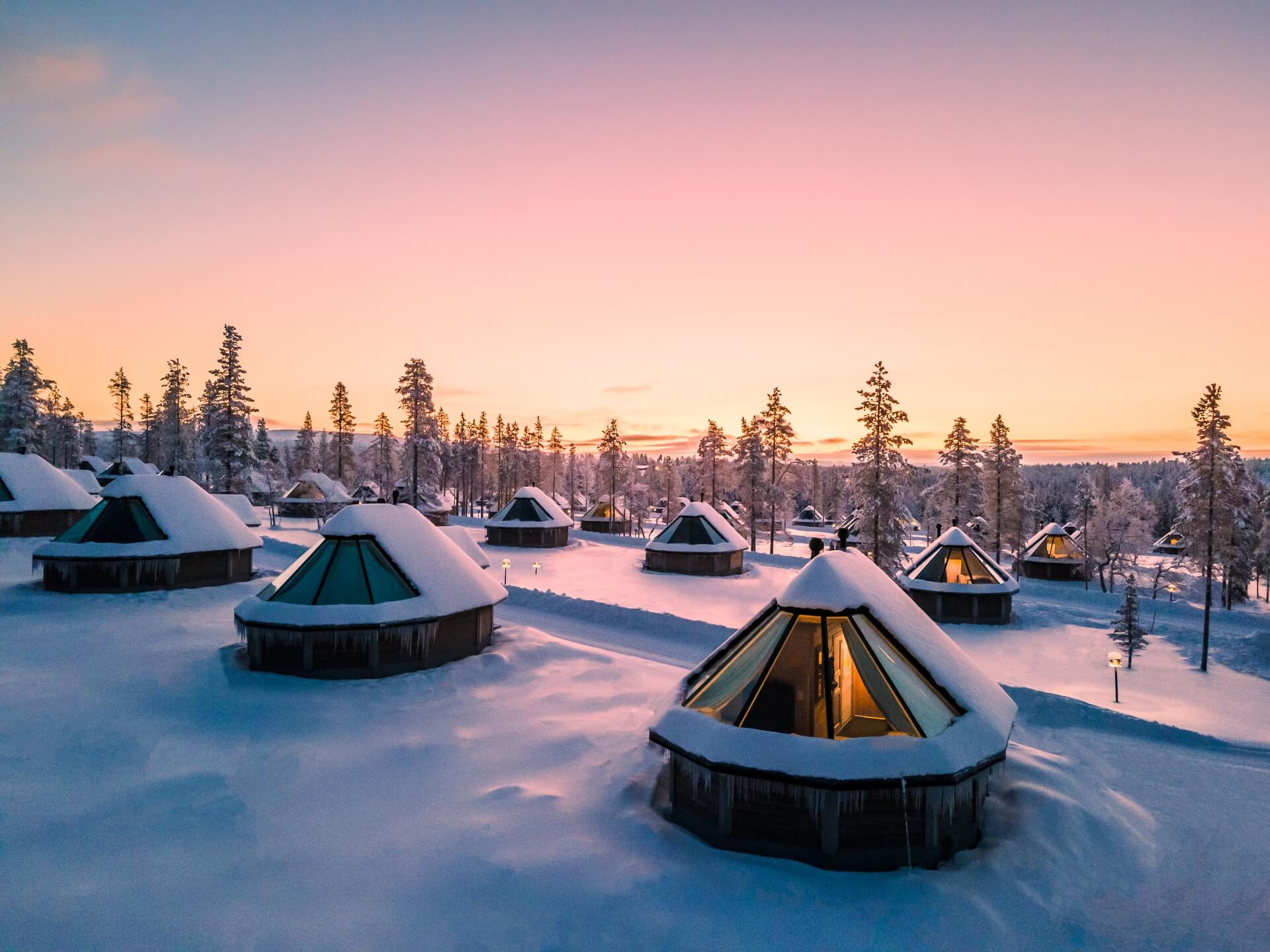 Snow-covered glass-roofed Aurora Cabins at sunset among pine trees
