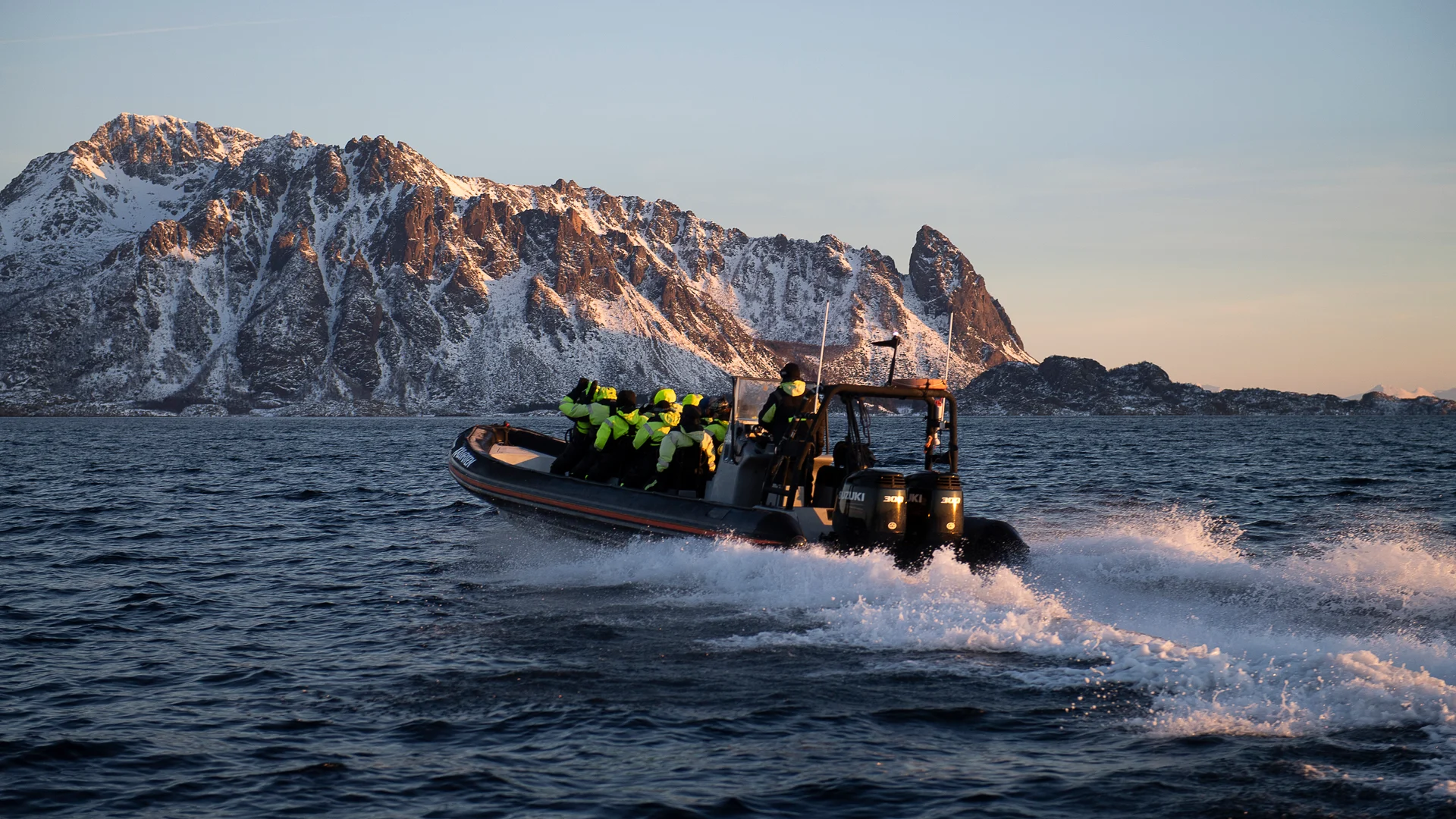 A boat of guests in yellow survival suits speeding past jagged snowy mountains